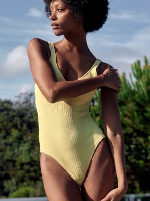 Woman in a white patterned dress standing outdoors in a sunlit street with a pink building in the background.