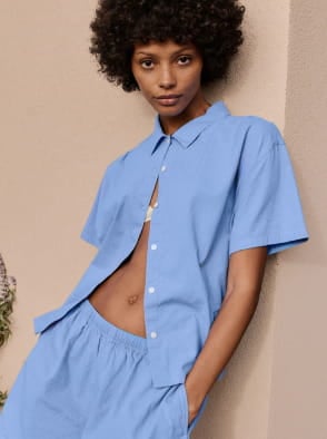 Woman in a white sleeveless top and beige pants standing on a sunlit stone pavement with an arched window in the background.