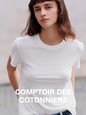 Woman in a white sleeveless top and beige pants standing on a sunlit stone pavement with an arched window in the background.