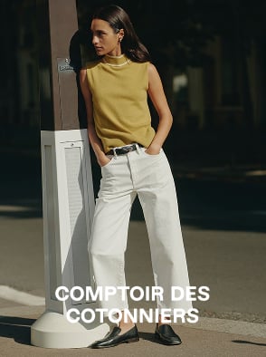 Woman in a white sleeveless top and beige pants standing on a sunlit stone pavement with an arched window in the background.