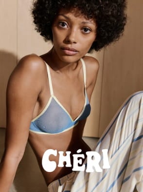 Woman in a white patterned dress standing outdoors in a sunlit street with a pink building in the background.