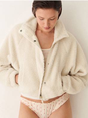 Woman in a white sleeveless top and beige pants standing on a sunlit stone pavement with an arched window in the background.