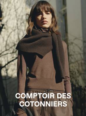 Woman in a white sleeveless top and beige pants standing on a sunlit stone pavement with an arched window in the background.